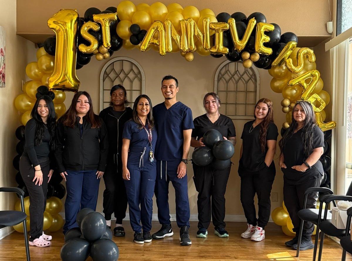 The Tooth Place team in Lynwood, CA: Dr. Alejandro Jimenez (center) and seven staff members in scrubs, smiling under a gold and black balloon arch that reads 1st Anniversary